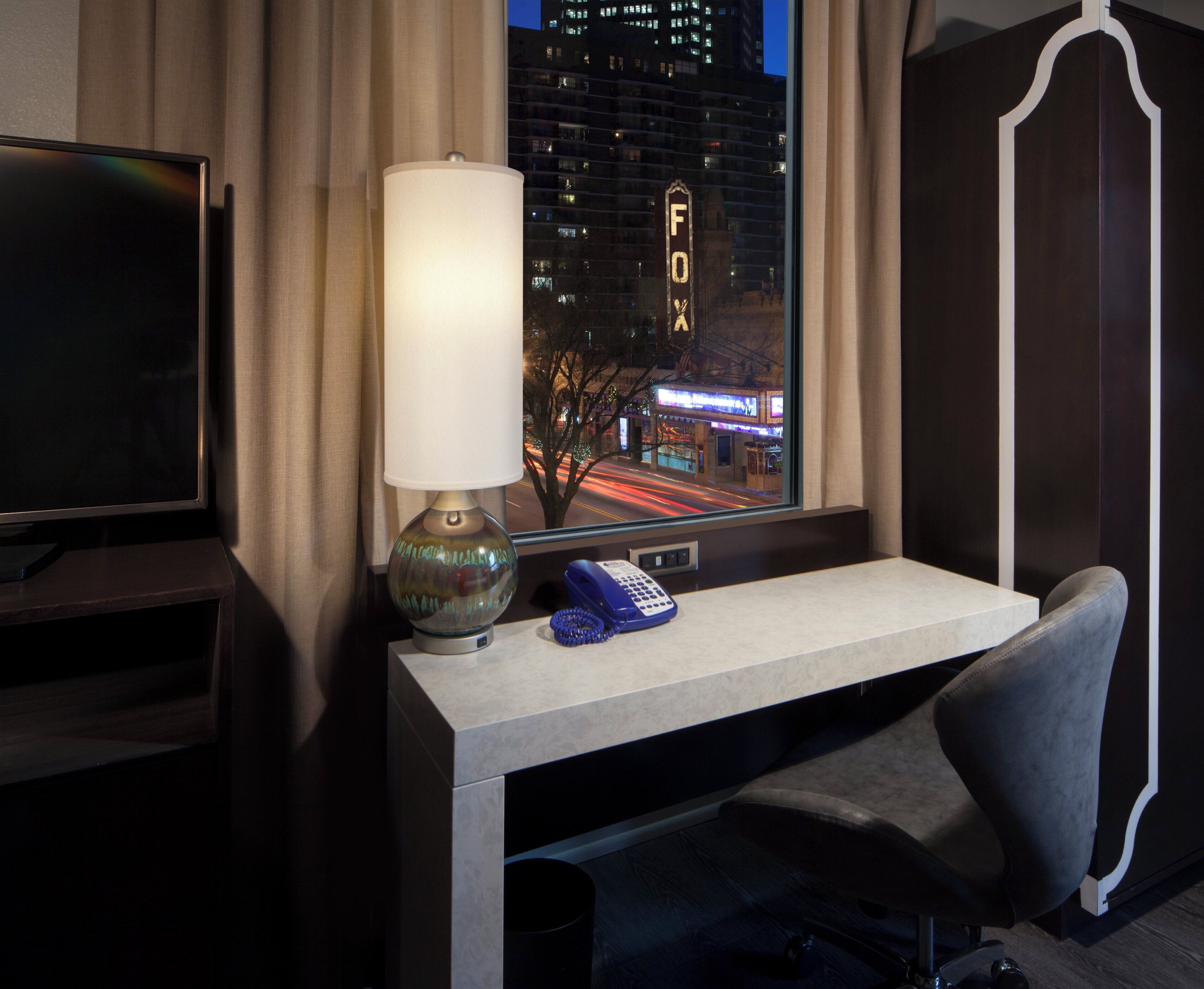 Een hotelkamer met een bureau, een lamp, een telefoon en uitzicht op een stadsbeeld 's nachts.