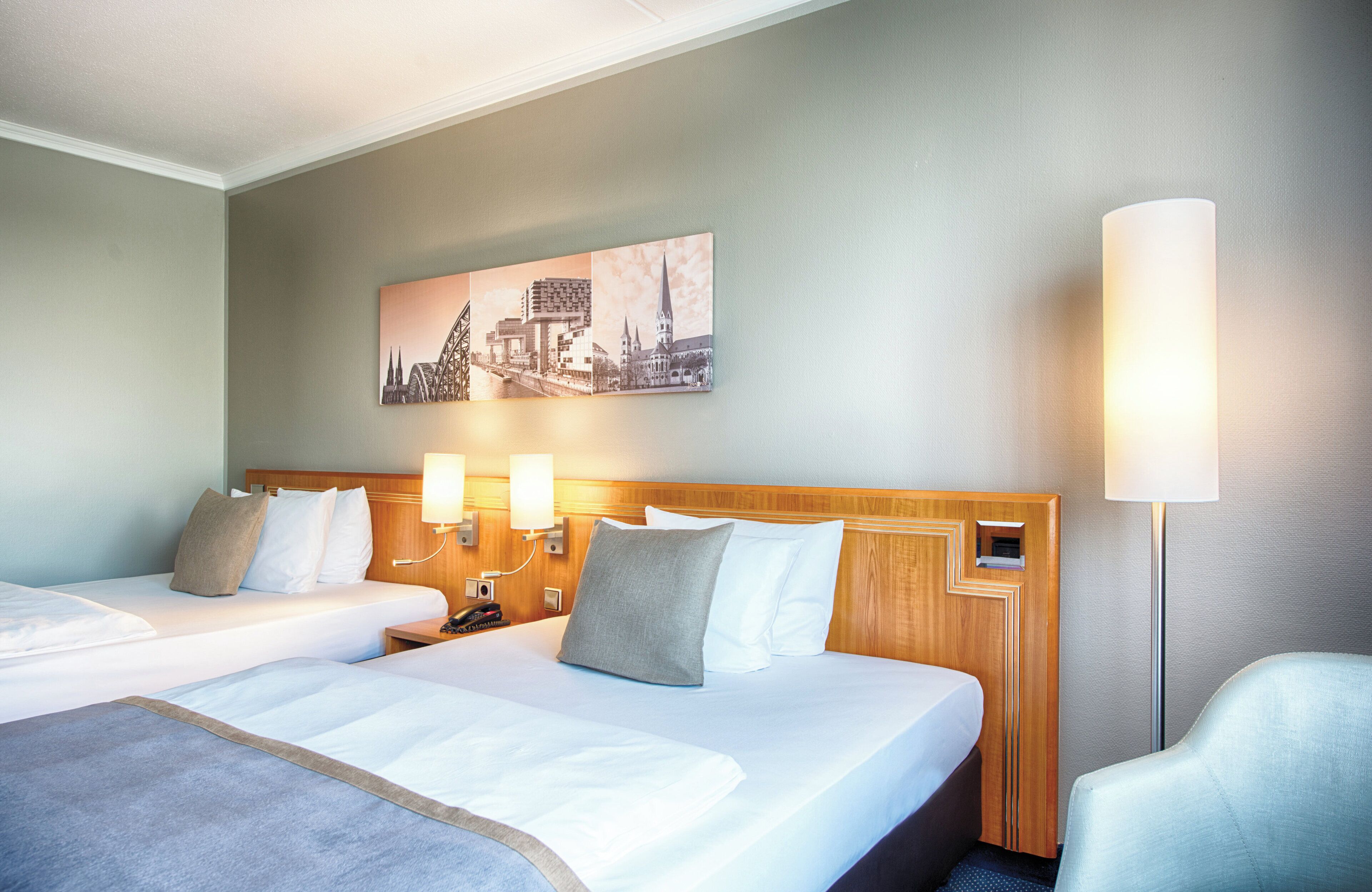 A hotel room with two beds, a headboard with a cityscape painting, bedside lamps, and a single chair.