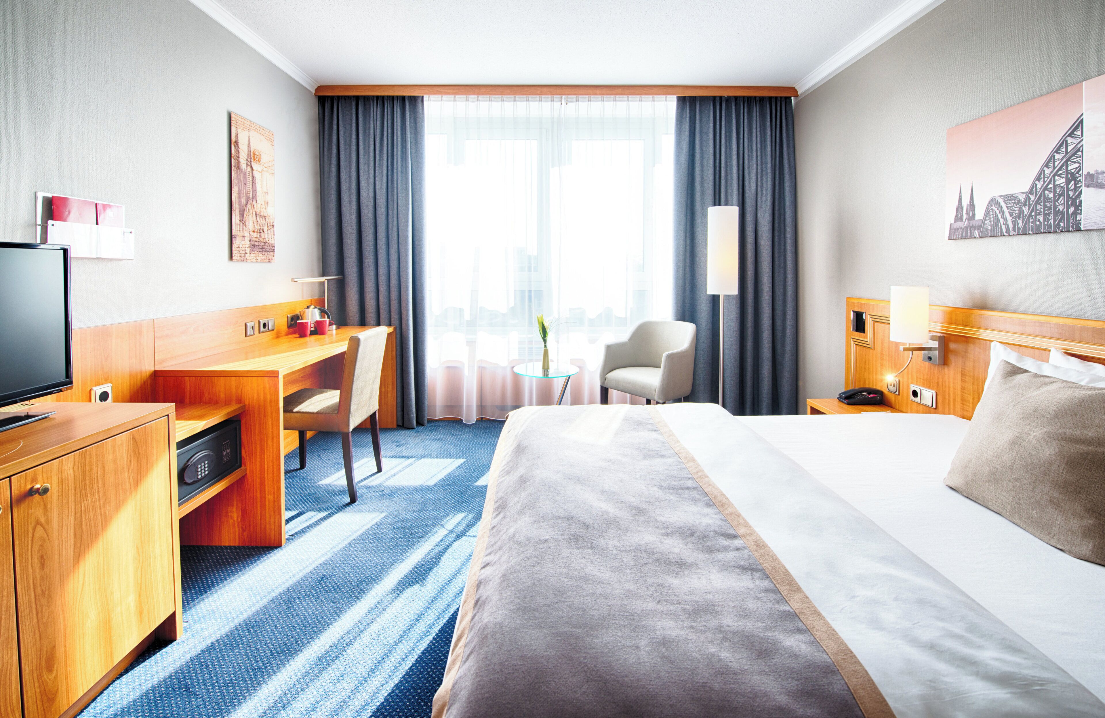 A hotel room with a large bed, a desk, a chair, a TV, and a window with curtains.