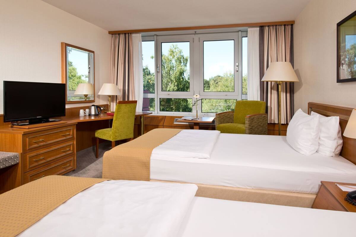 A hotel room with two beds, a desk with a TV, a chair, a lamp, and a window with a view of trees.
