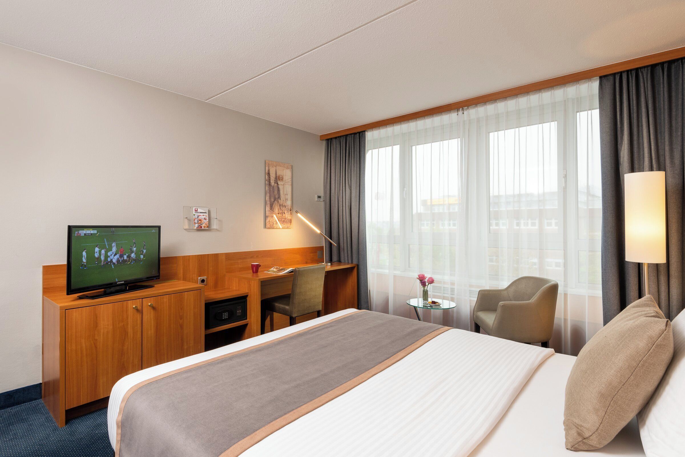 Ein Hotelzimmer mit Bett, Schreibtisch, Fernseher, Stuhl und einem Fenster mit Vorhängen.