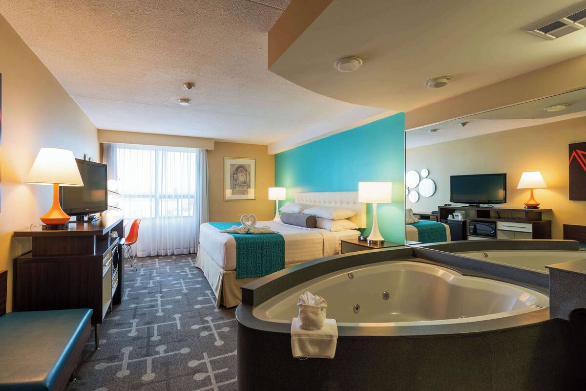 Une chambre d’hôtel dotée d’un grand lit, d’un coin salon avec un bureau et une chaise, d’une télévision, d’une salle de bain avec baignoire et d’une grande fenêtre avec des rideaux.
