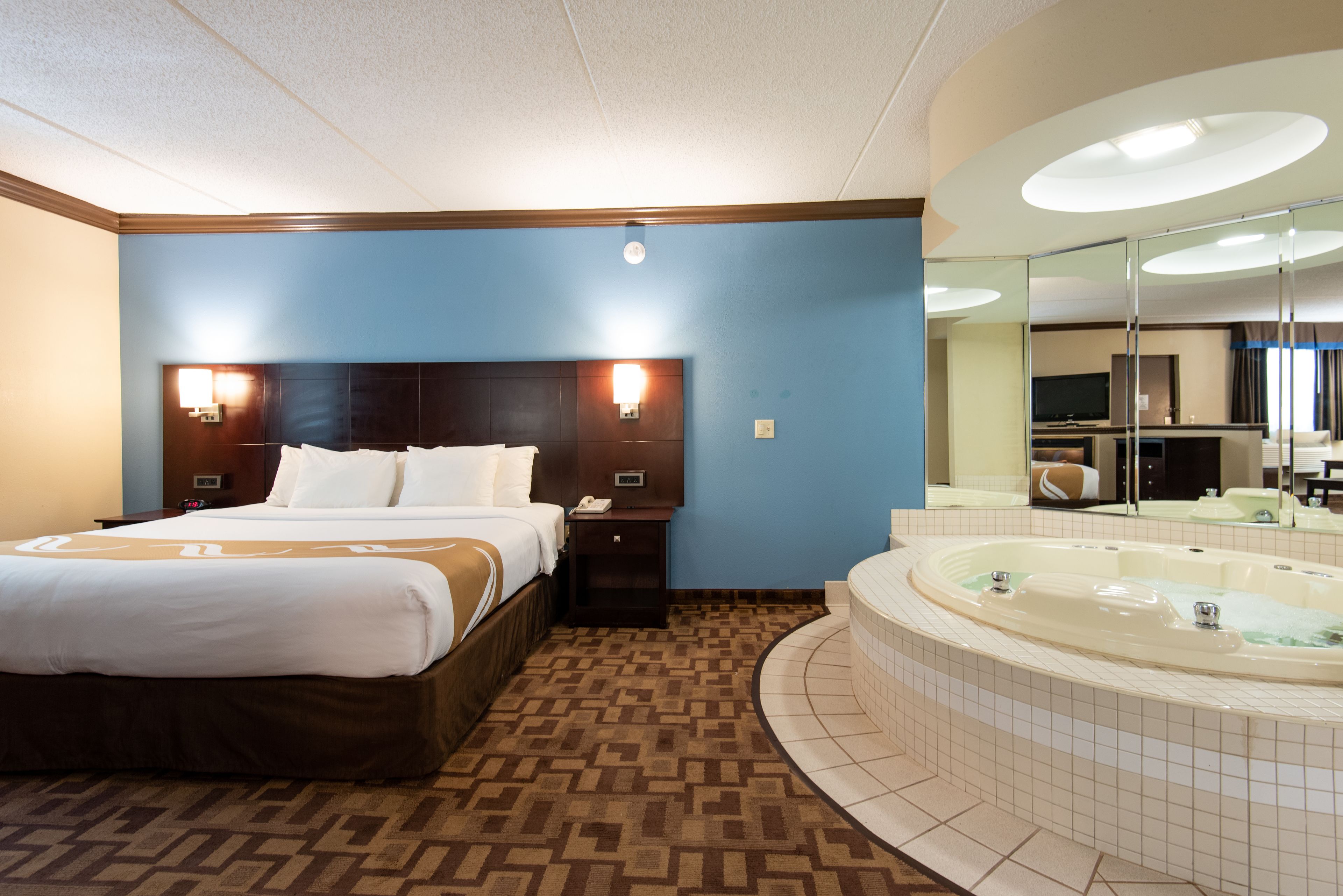 A hotel room with a large bed, a separate sofa bed, a built-in TV, a large mirror, and a separate bathroom with a bathtub.
