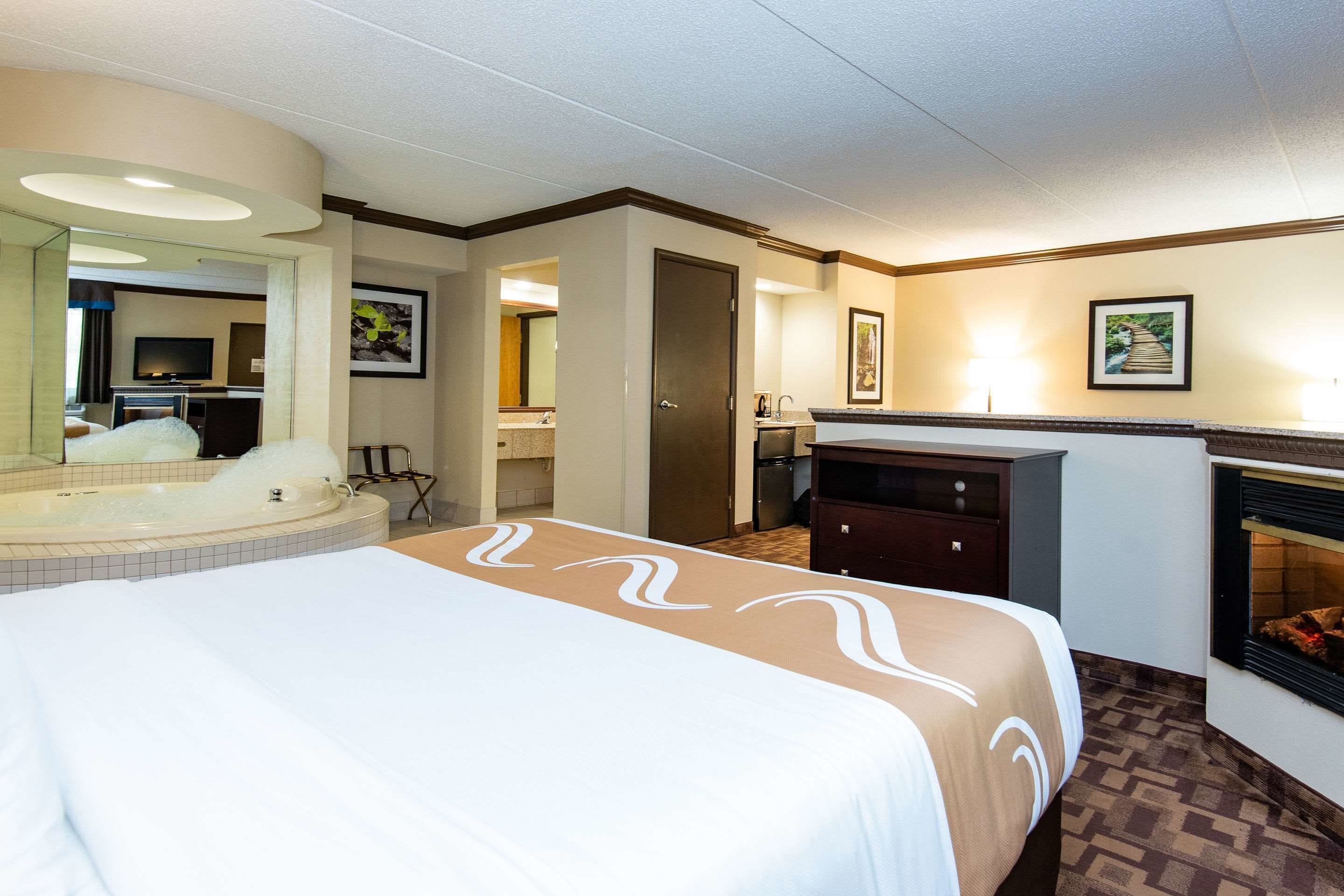 A hotel room with a large bed, a bathroom with a bathtub, a desk, a fireplace, and a television.