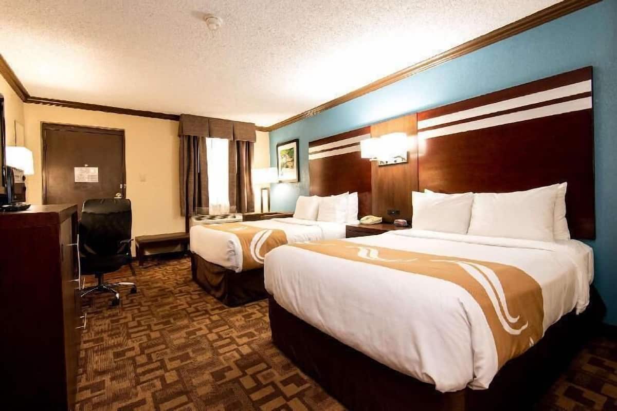 A hotel room with two beds, a desk, a chair, and a door.
