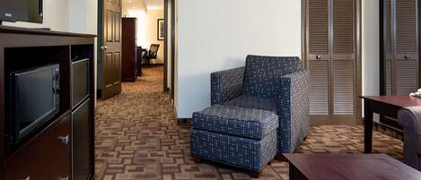 A hotel room with a patterned carpet, a blue upholstered chair, a wooden table, and a TV on a cabinet.