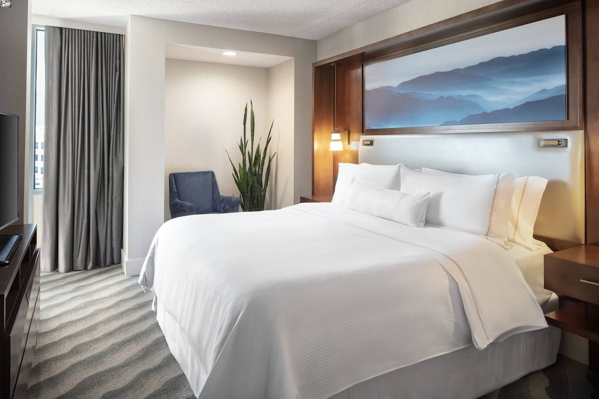 A hotel room with a large bed, a desk, a chair, a potted plant, and a painting on the wall.