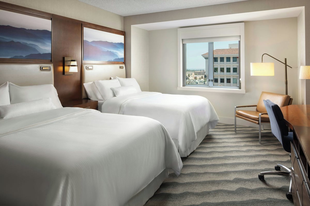 A hotel room with two beds, a desk, a chair, a lamp, and a window with a city view.