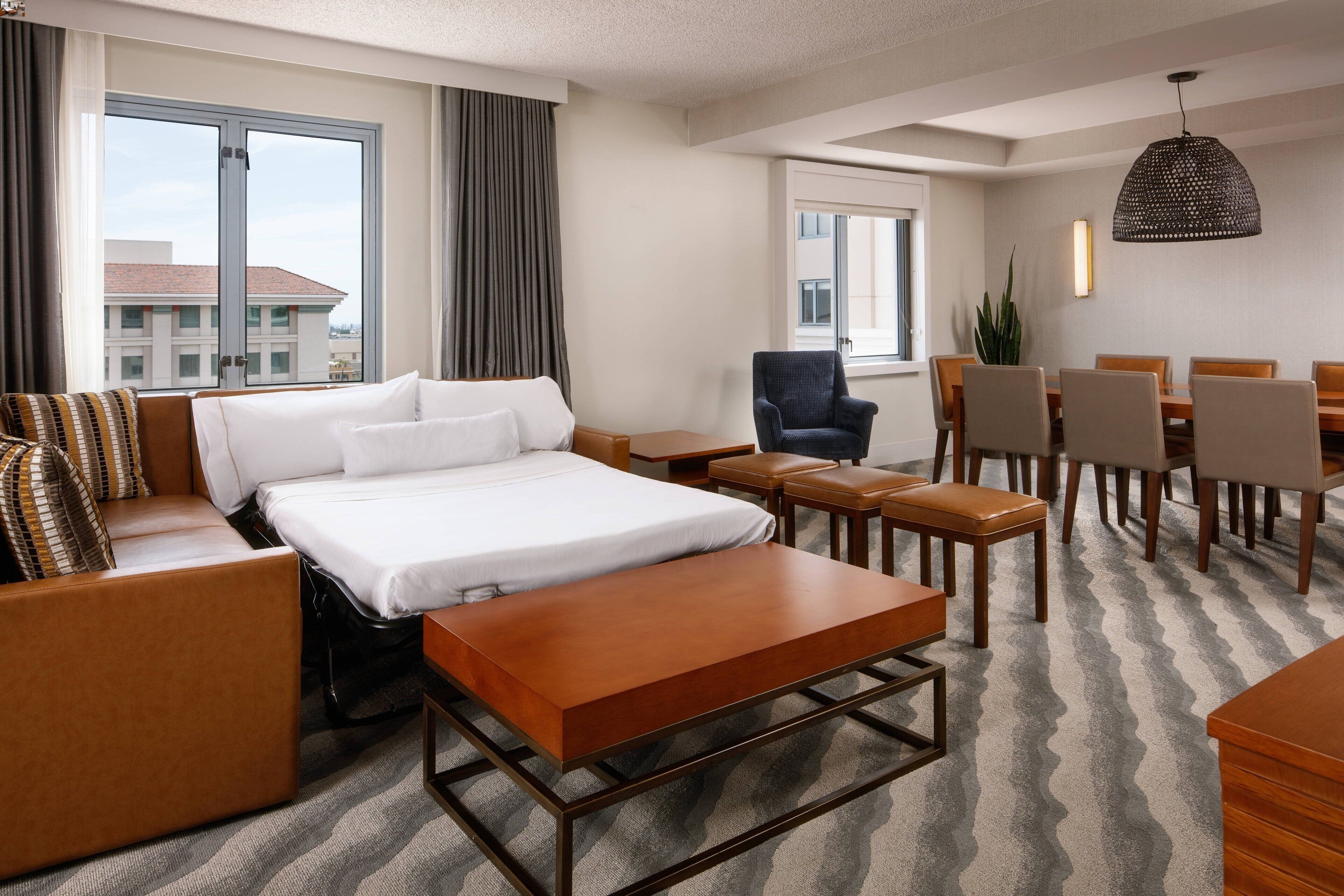 Habitación de hotel con cama, sofá, mesa de comedor con sillas, una silla y lámpara colgante.