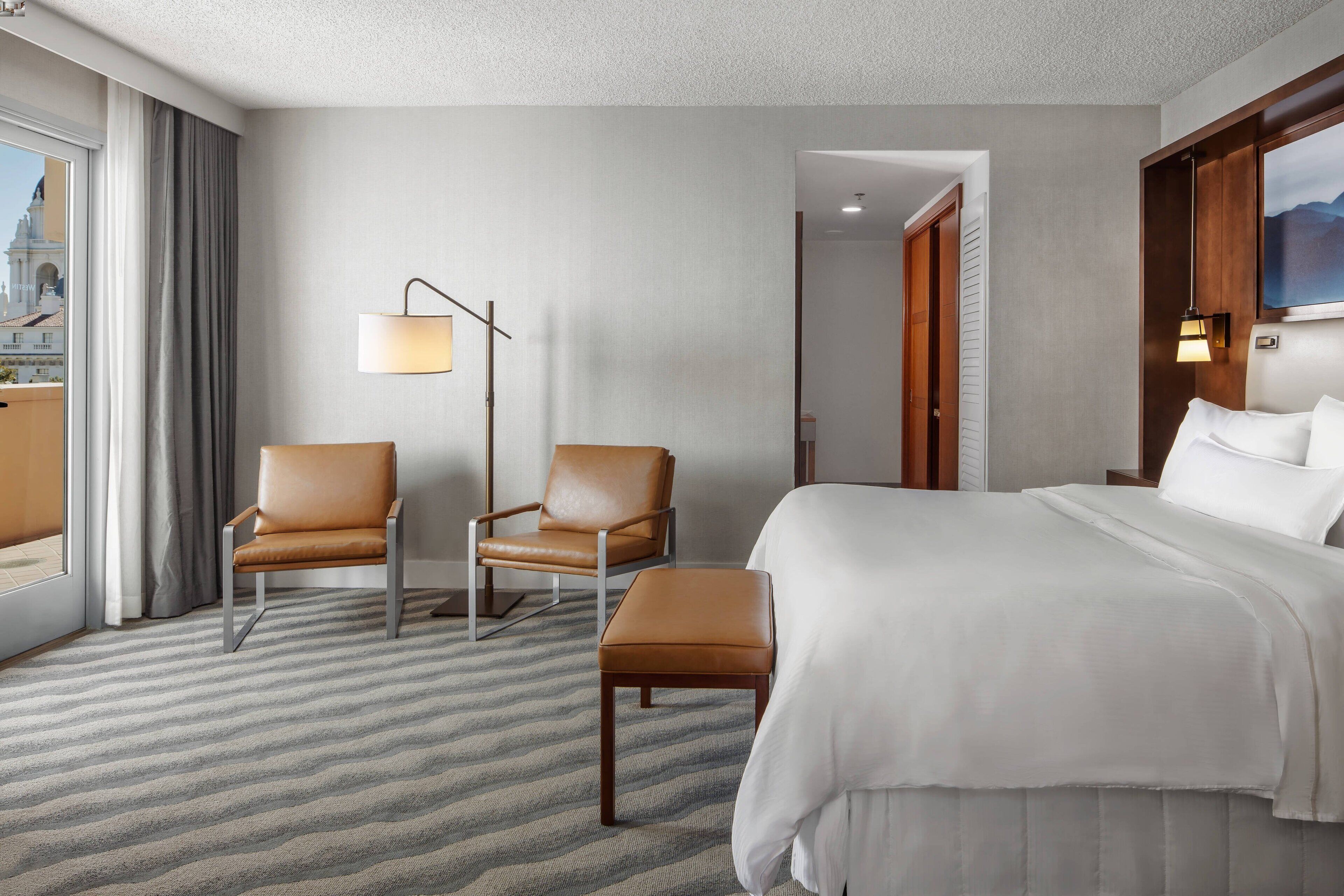 Quarto de hotel com uma cama grande, duas cadeiras, um abajur e vista para a cidade através da janela.