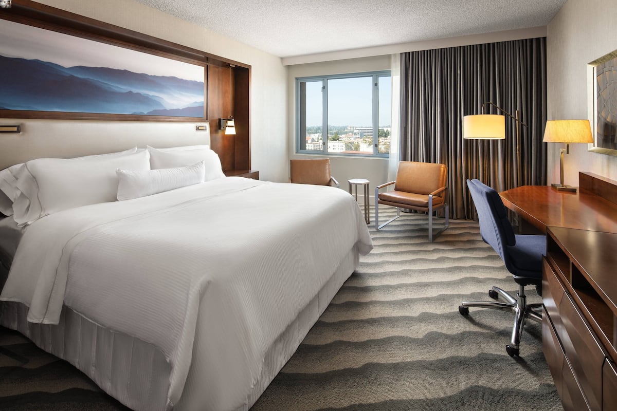 A hotel room with a large bed, a desk, a chair, a small table, and a view of the city through a window.