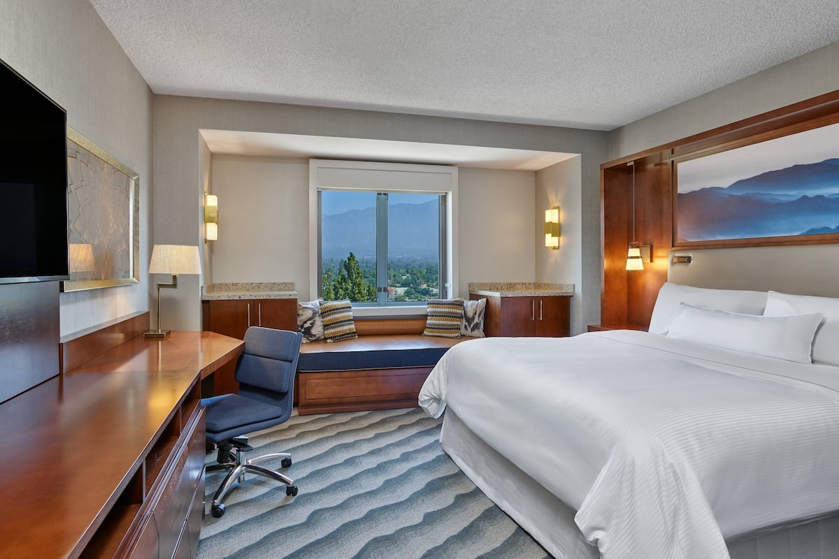 A hotel room with a large bed, a desk with a chair, a view of mountains through a window, and a mounted TV.
