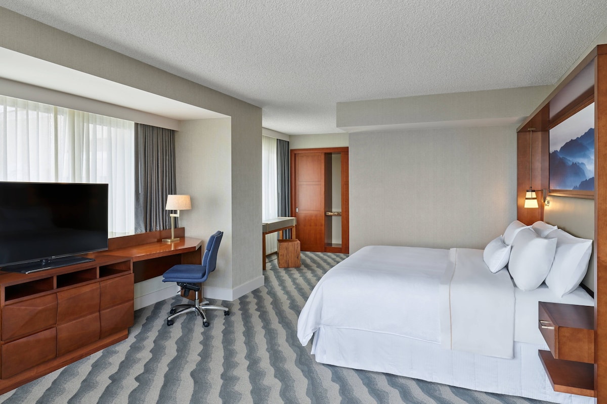 A hotel room with a large bed, a desk with a chair, a television, and a bathroom visible through an open door.