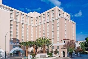 Un hotel grande de varios pisos con una entrada destacada, palmeras y un cielo azul claro.