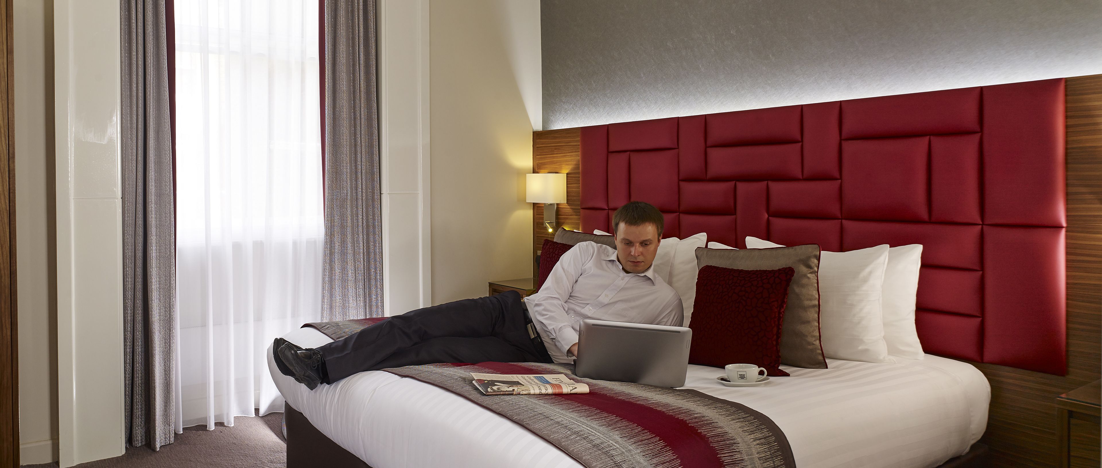 A hotel room with a bed, a laptop, a cup, and a newspaper.