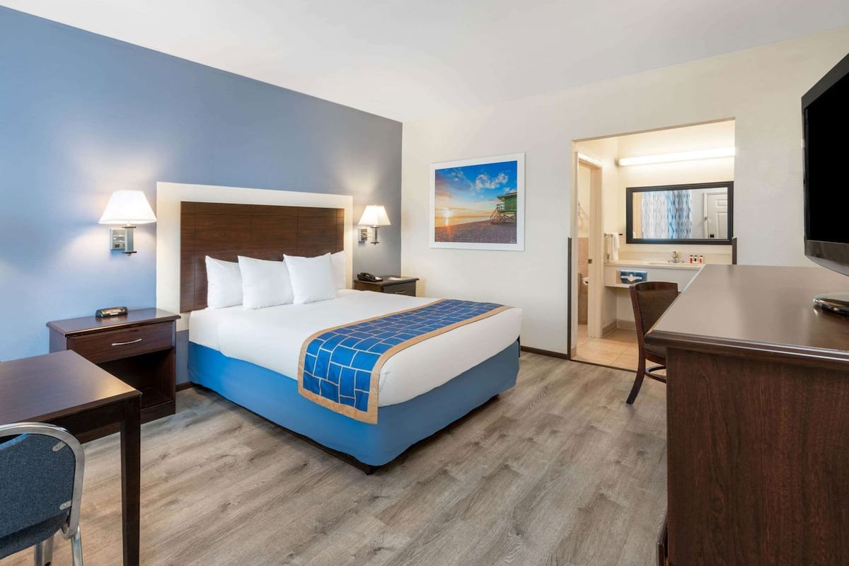 A hotel room with a bed, bedside tables, a desk, a chair, a television, and a bathroom visible through an open door.