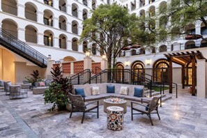Une cour spacieuse dotée d’un espace de détente central comprenant un canapé et des fauteuils, le tout entouré d’un sol carrelé et d’un grand arbre.