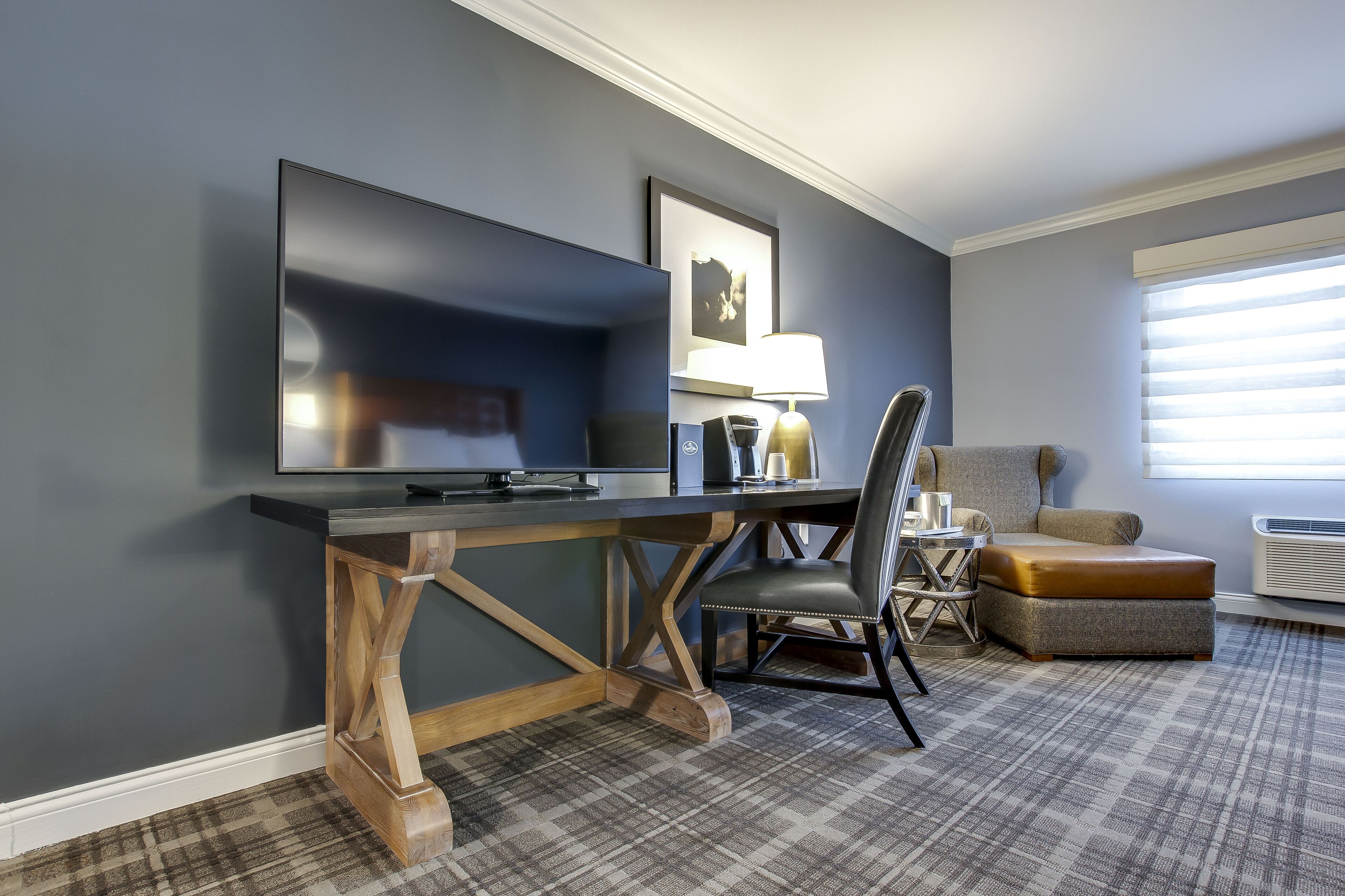 Habitación de hotel con televisor de pantalla plana grande, escritorio de madera, silla, otomana pequeña y ventana con persianas.