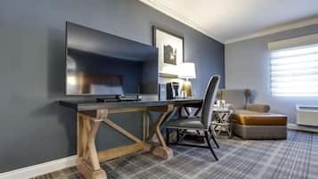 Habitación de hotel con televisor de pantalla plana grande, escritorio de madera, silla, otomana pequeña y ventana con persianas.