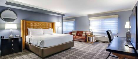 A hotel room with a large bed, a desk, a chair, a sofa, a round mirror, and a window with blinds.