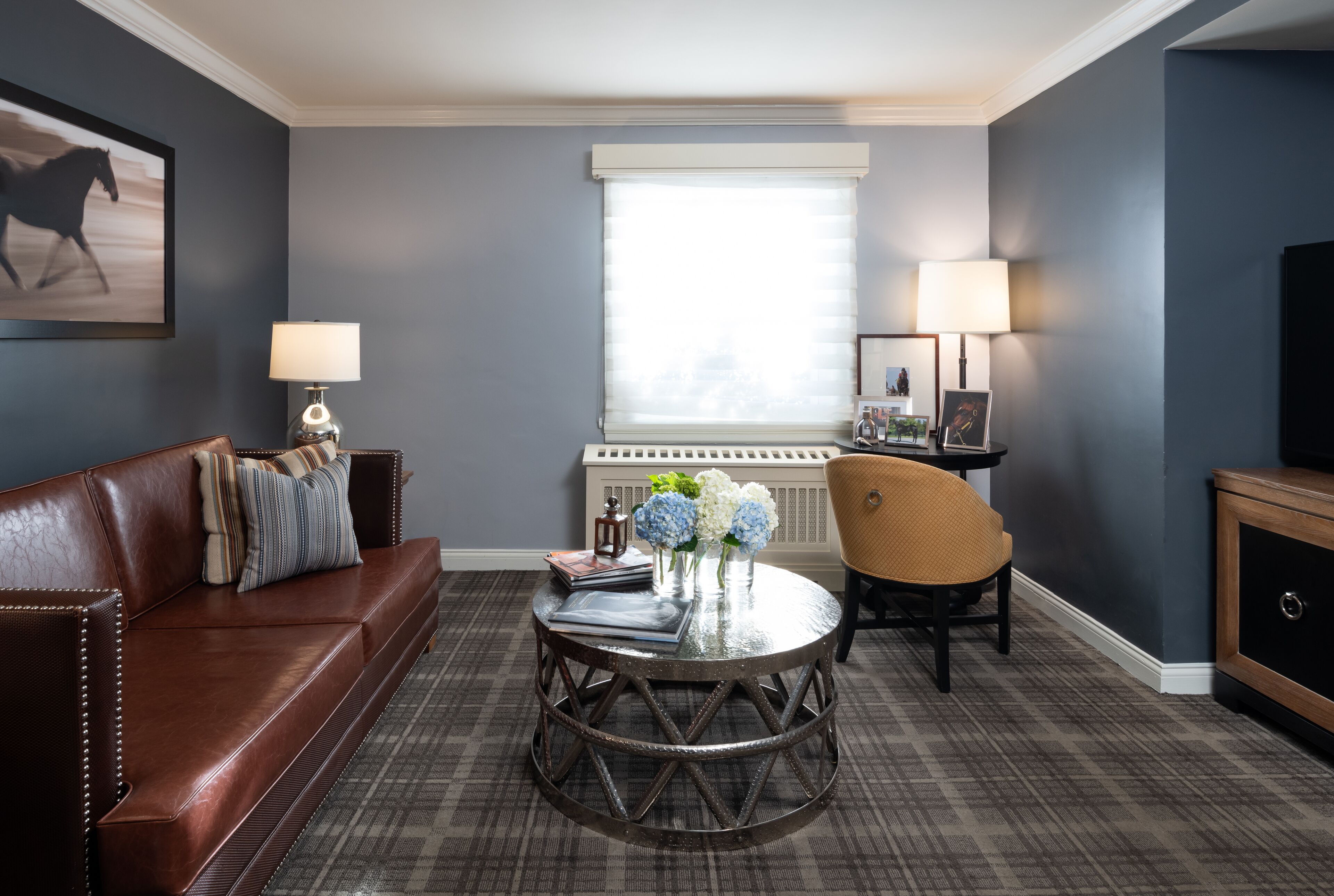 A modern living room with a brown leather sofa, a round coffee table with decorative items, a wicker chair, and a large window with blinds.