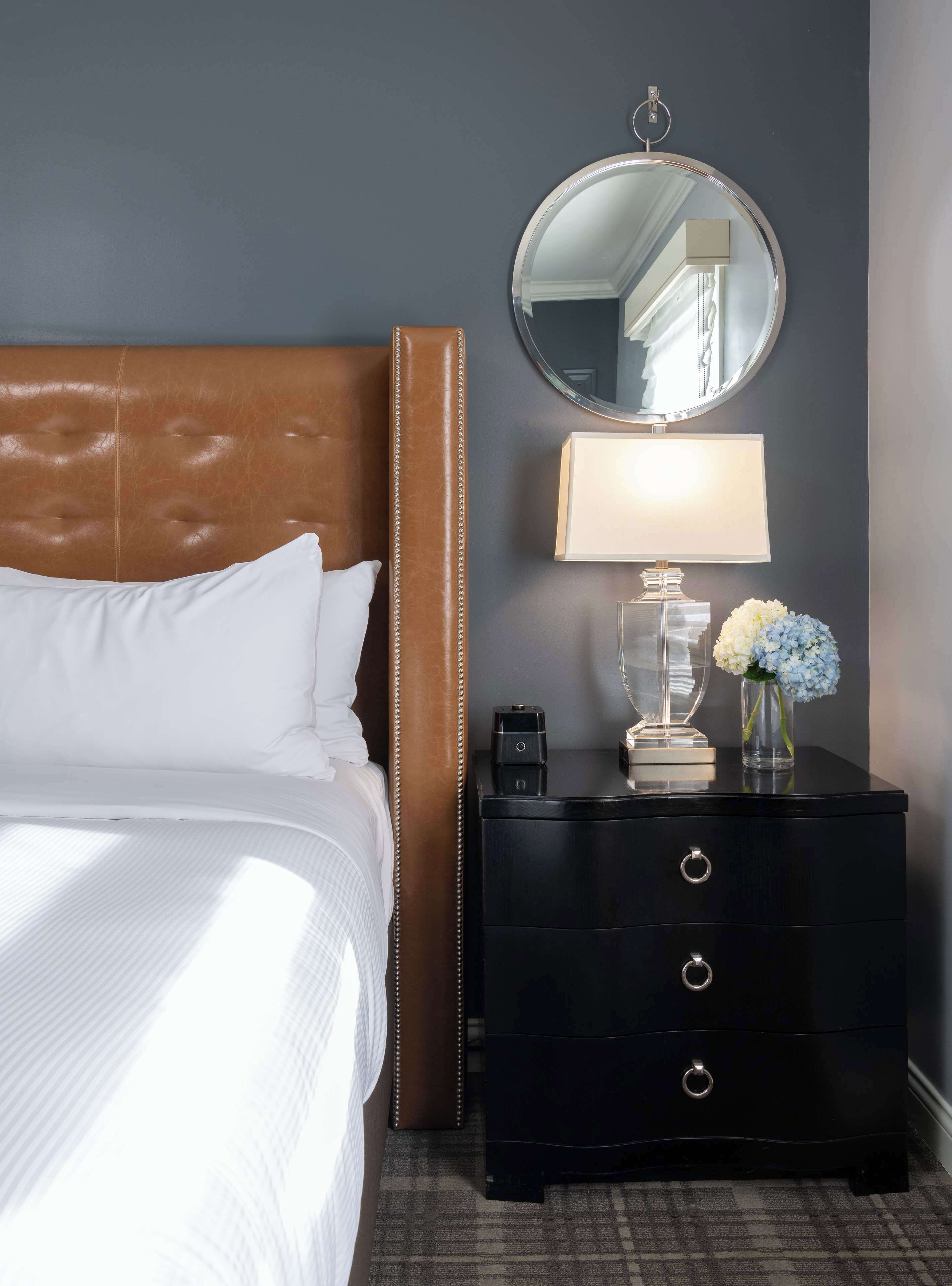 A neatly made bed with white linens, a brown leather headboard, a black nightstand with a mirror, a lamp, and a vase with flowers.