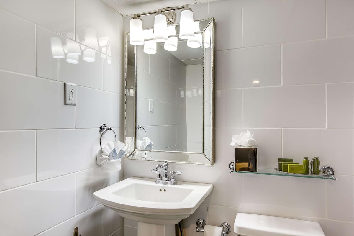 Un baño con lavamanos blanco, espejo e inodoro. Se observa un perchero con una toalla y una caja de pañuelos en la pared.