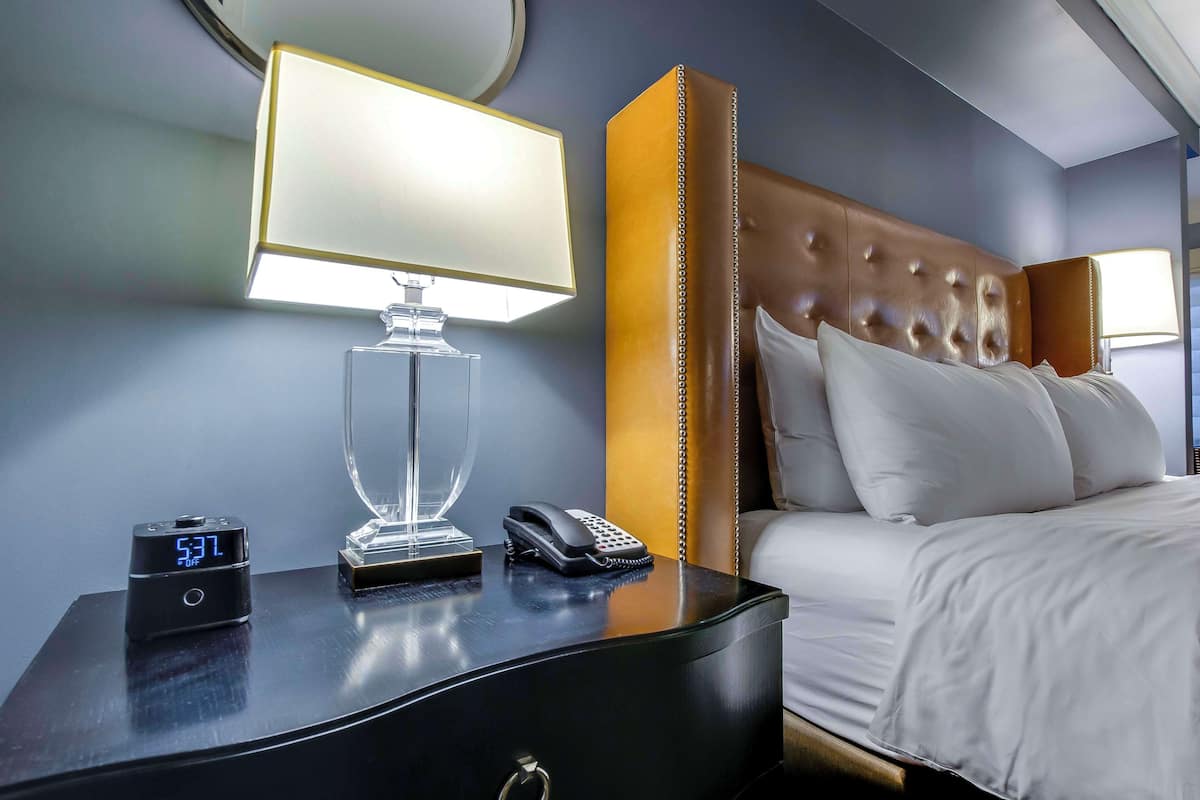 Habitación de hotel con cama, mesita de noche con lámpara, reloj digital y teléfono.