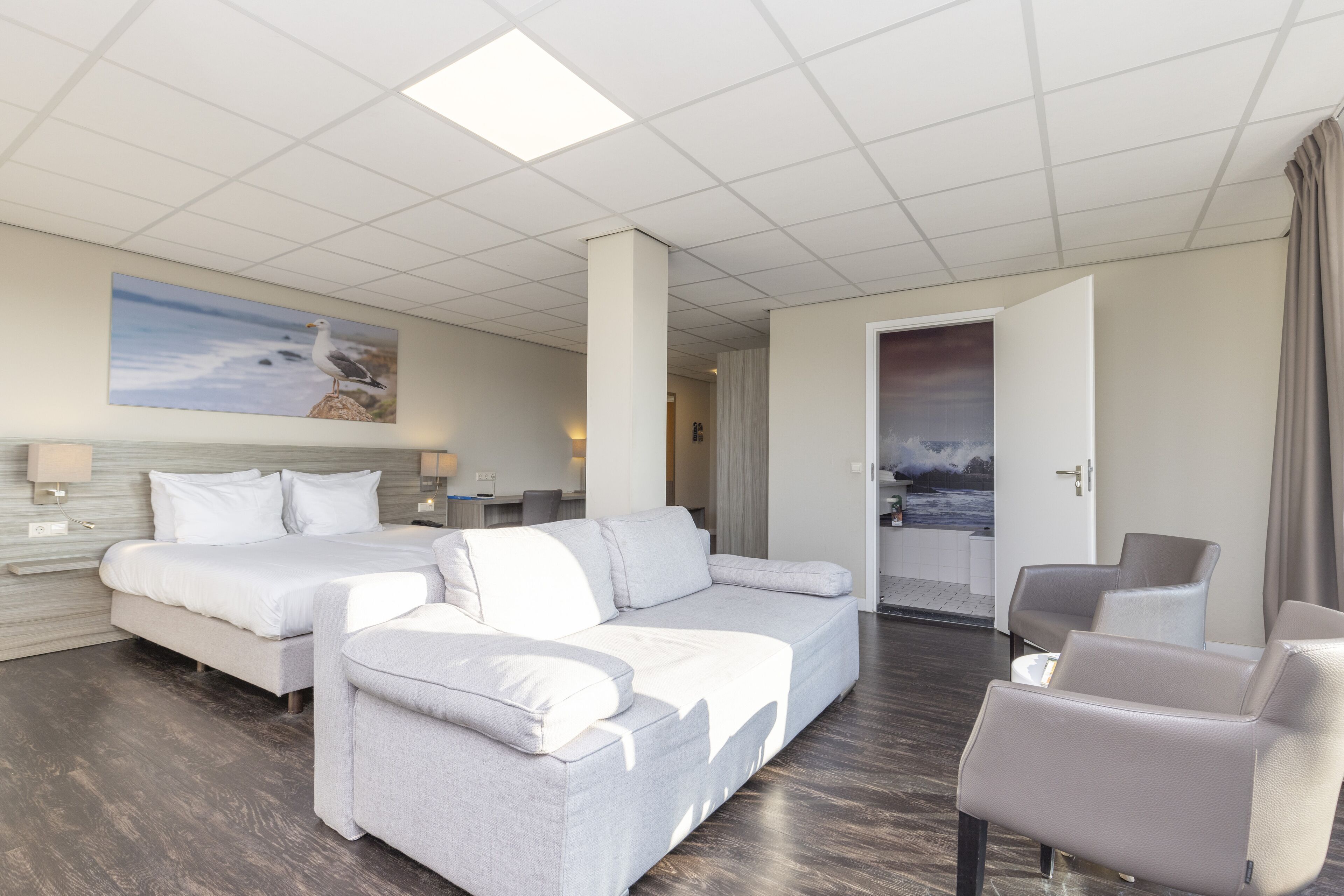 A modern hotel room with a large bed, a sofa, and a chair. There is a painting on the wall and a view of the sea through the window.