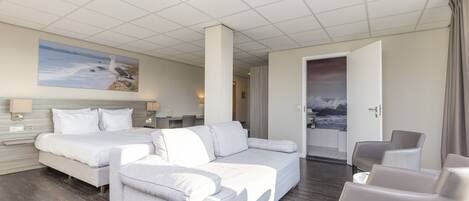 A modern hotel room with a large bed, a sofa, and a chair. There is a painting on the wall and a view of the sea through the window.