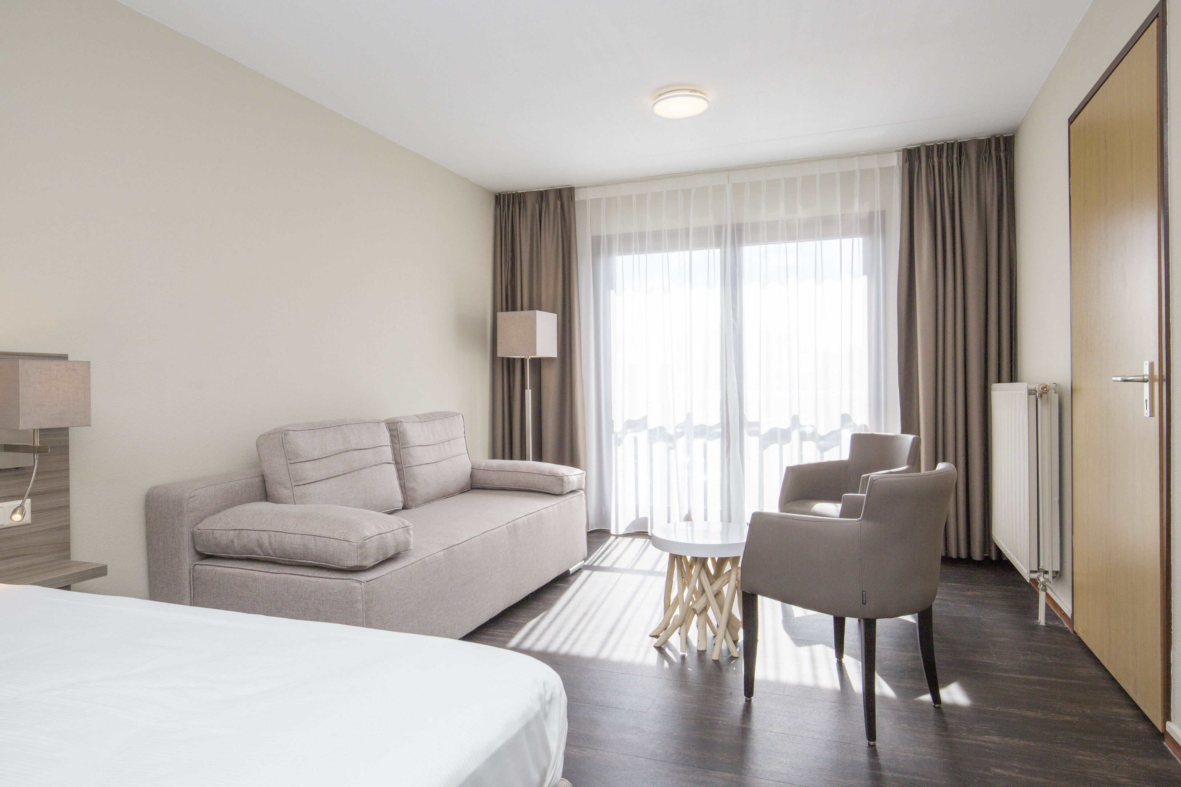 A hotel room with a bed, a sofa, a dining table with chairs, a lamp, and a large window with curtains.