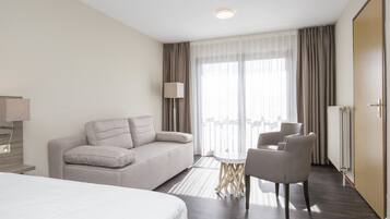 Quarto de hotel com cama, sofá, mesa de jantar com cadeiras, luminária e uma janela grande com cortinas.