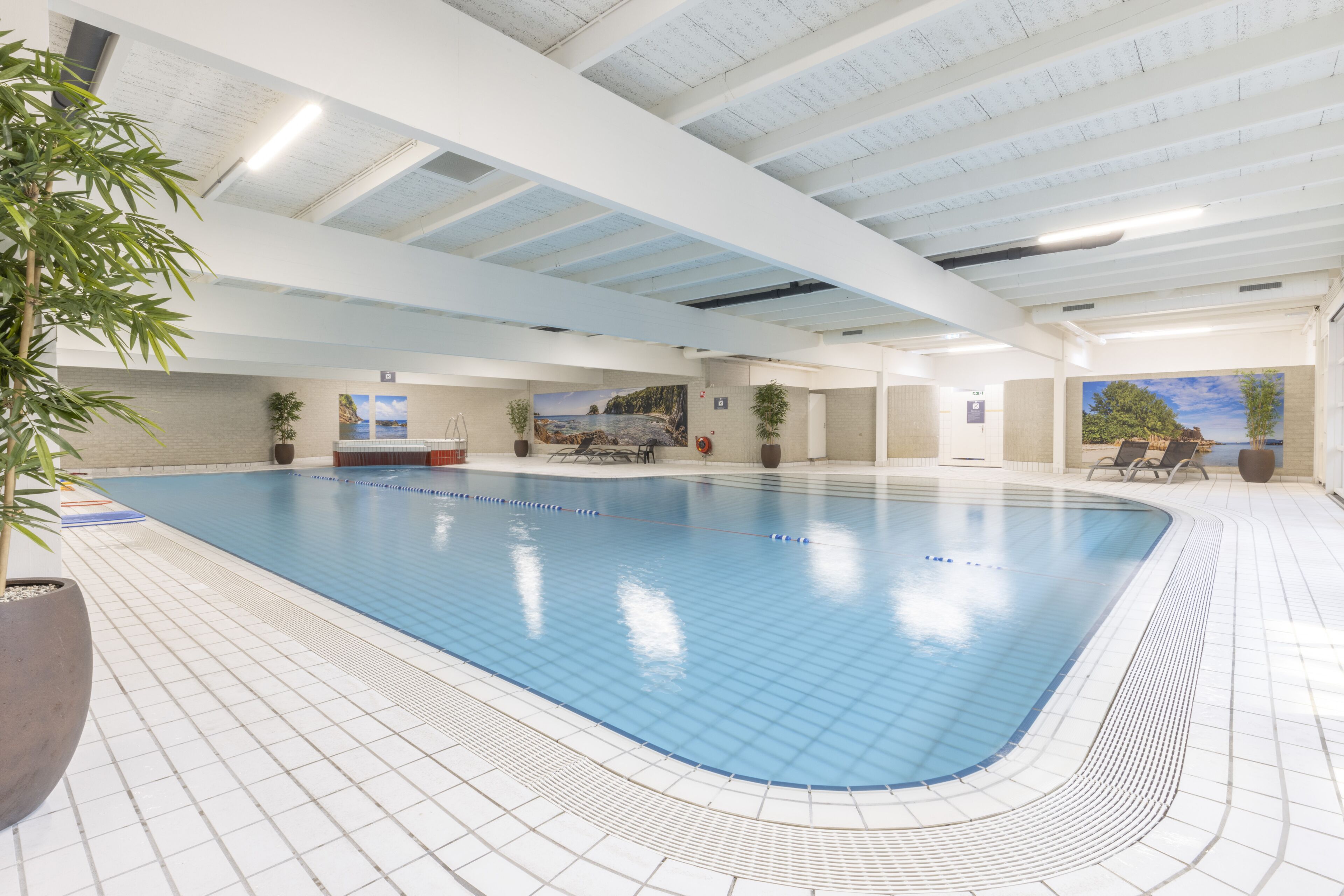 A large indoor swimming pool with a tiled floor, ceiling with exposed beams, and potted plants around the pool area.