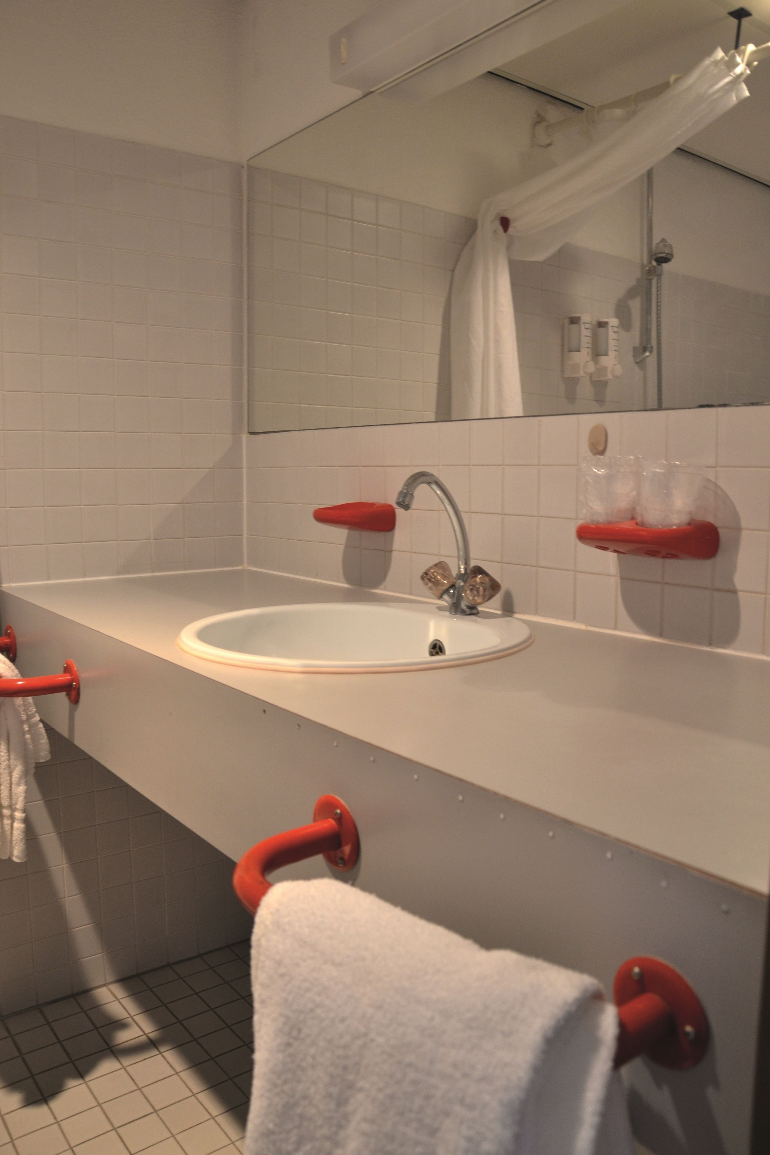 Un baño con lavamanos blanco, sujetabandejas rojos, un espejo y cortina de ducha.
