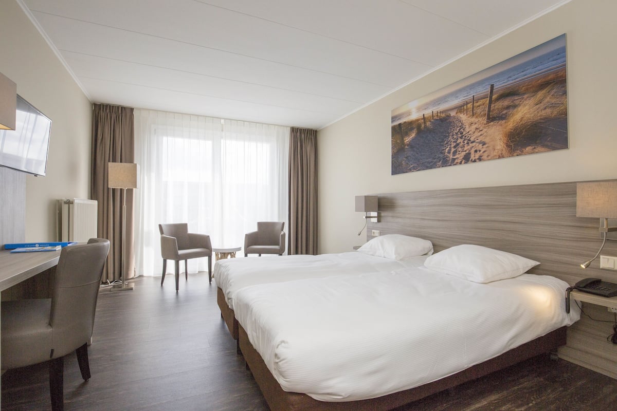 A hotel room with a large bed, two bedside tables, a desk with a chair, a TV, and a painting on the wall.