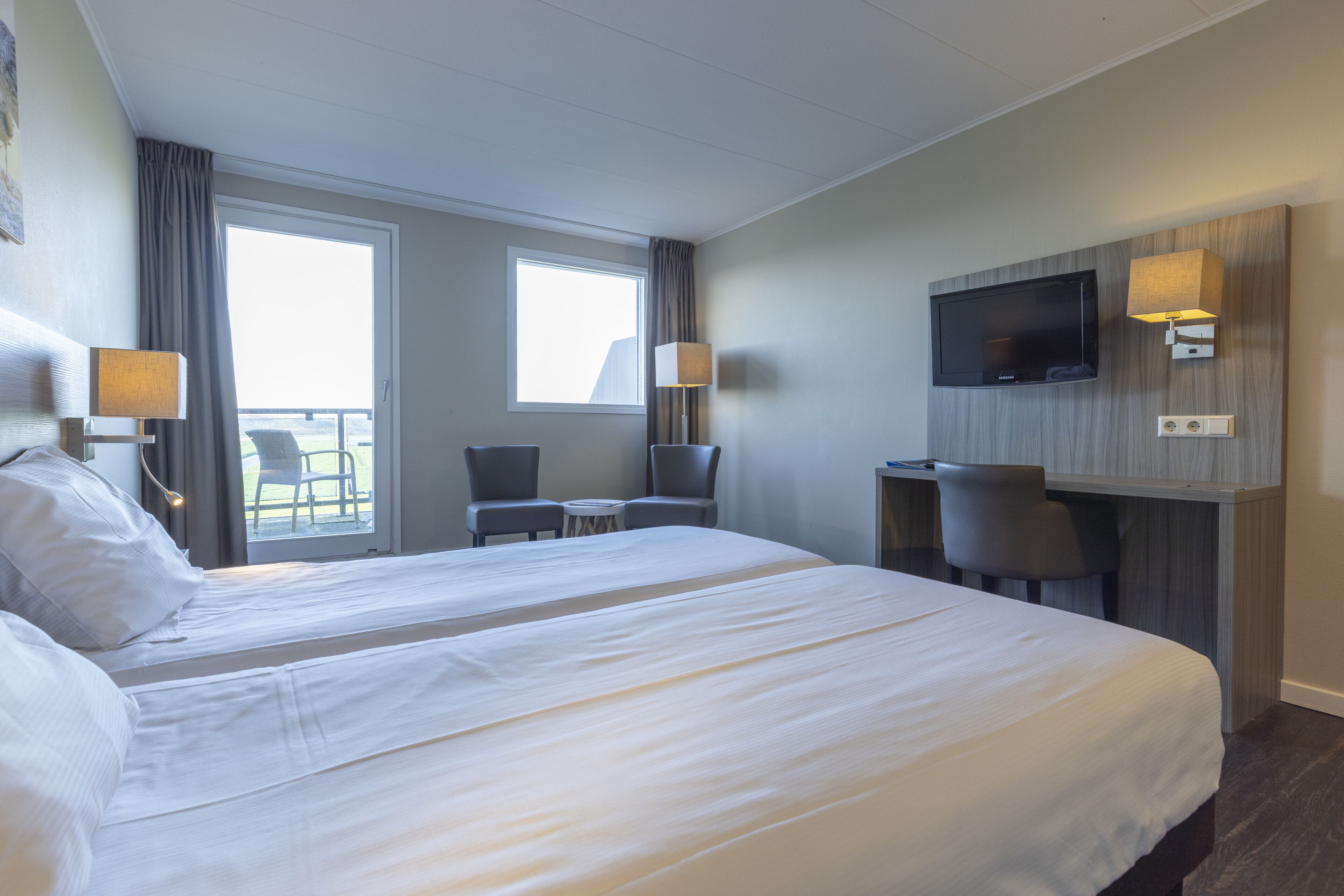 A hotel room with two beds, a TV mounted on the wall, and a balcony with chairs.
