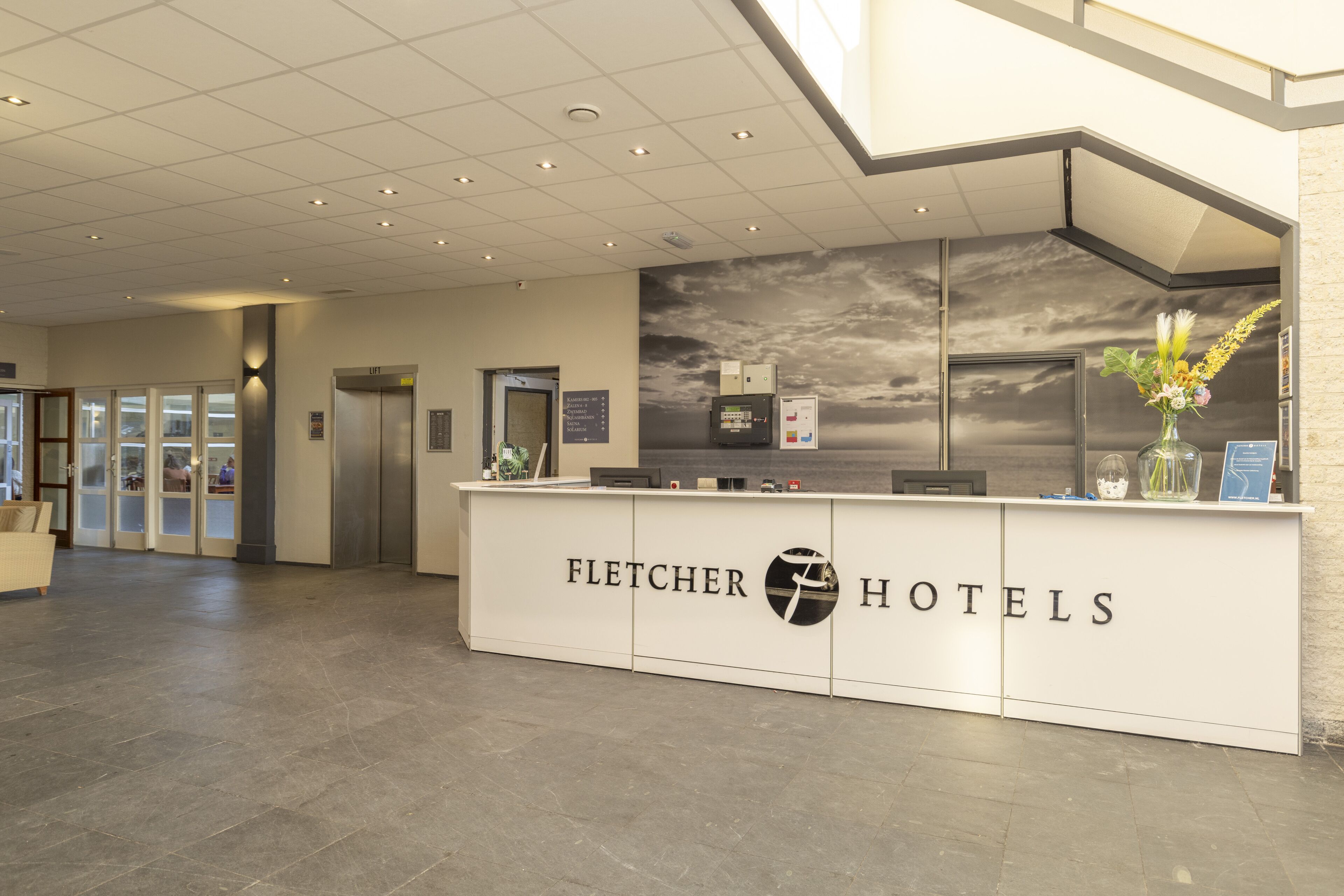 A reception desk with the Fletcher Hotels logo, a modern interior with a tiled floor, and a large window with a view of the sky.