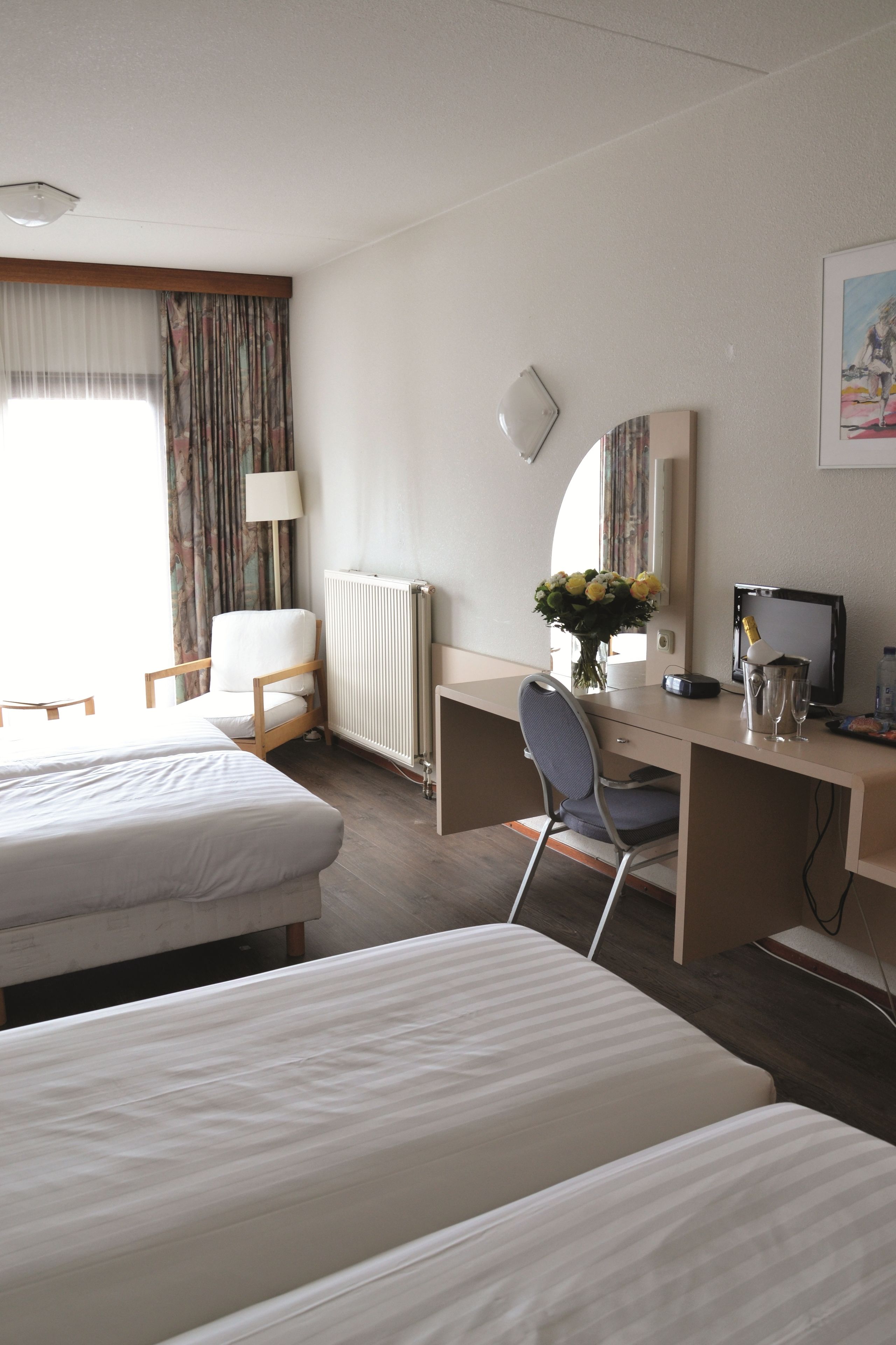 Une chambre d’hôtel avec deux lits, un bureau avec un ordinateur, une chaise, une lampe, un radiateur et une fenêtre avec des rideaux.