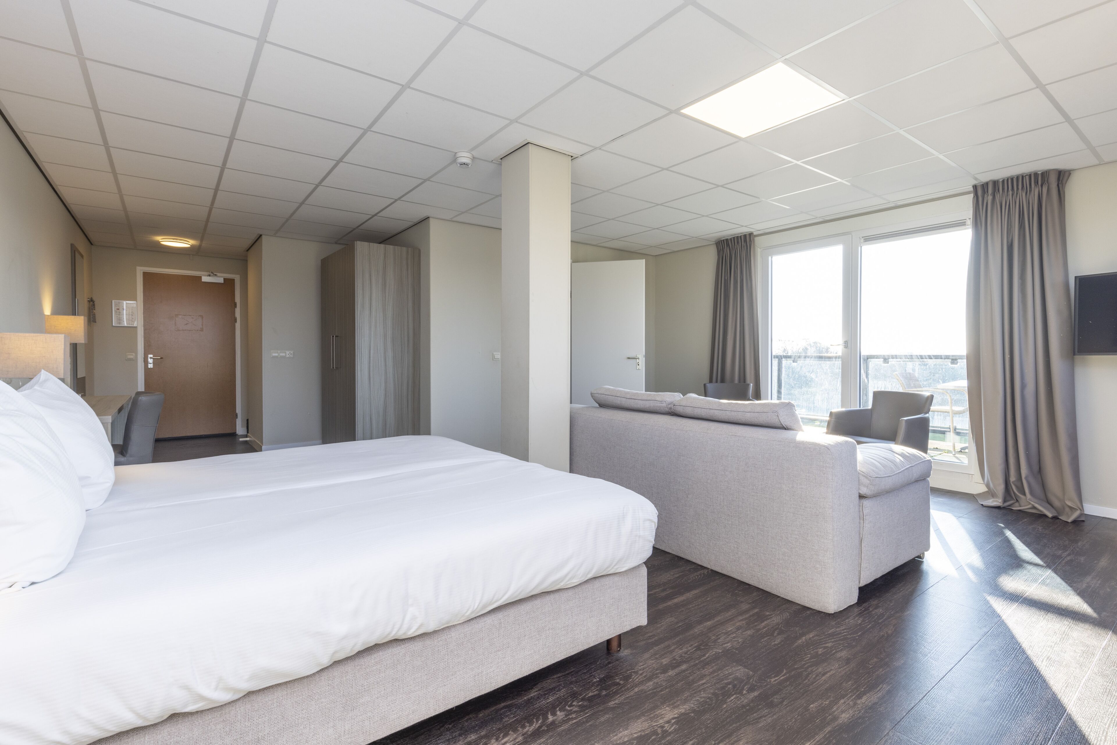 A hotel room with a large bed, a sofa, a desk, and a view of the city through large windows.