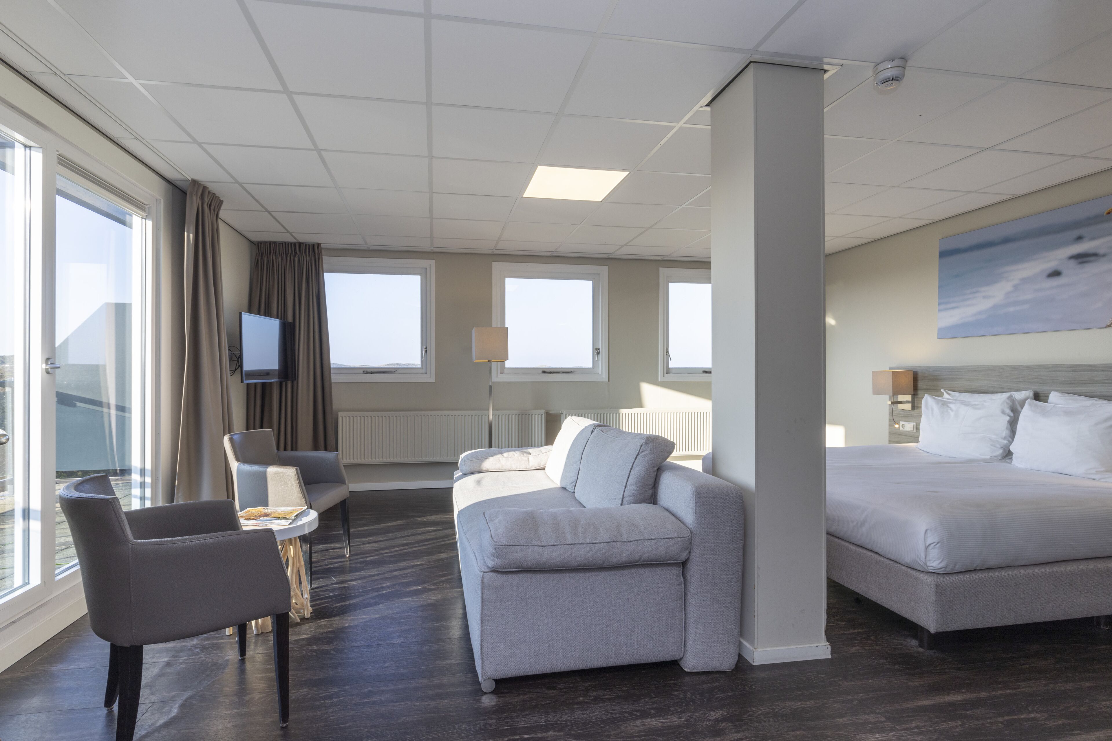 Habitación de hotel moderna con un sofá gris, una cama, un televisor y ventanas amplias que ofrecen una vista.