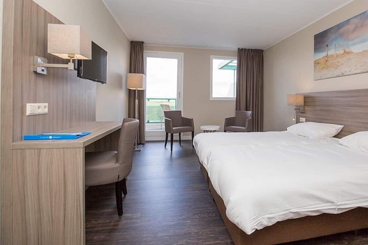 A hotel room with a bed, desk, chairs, and a painting on the wall.