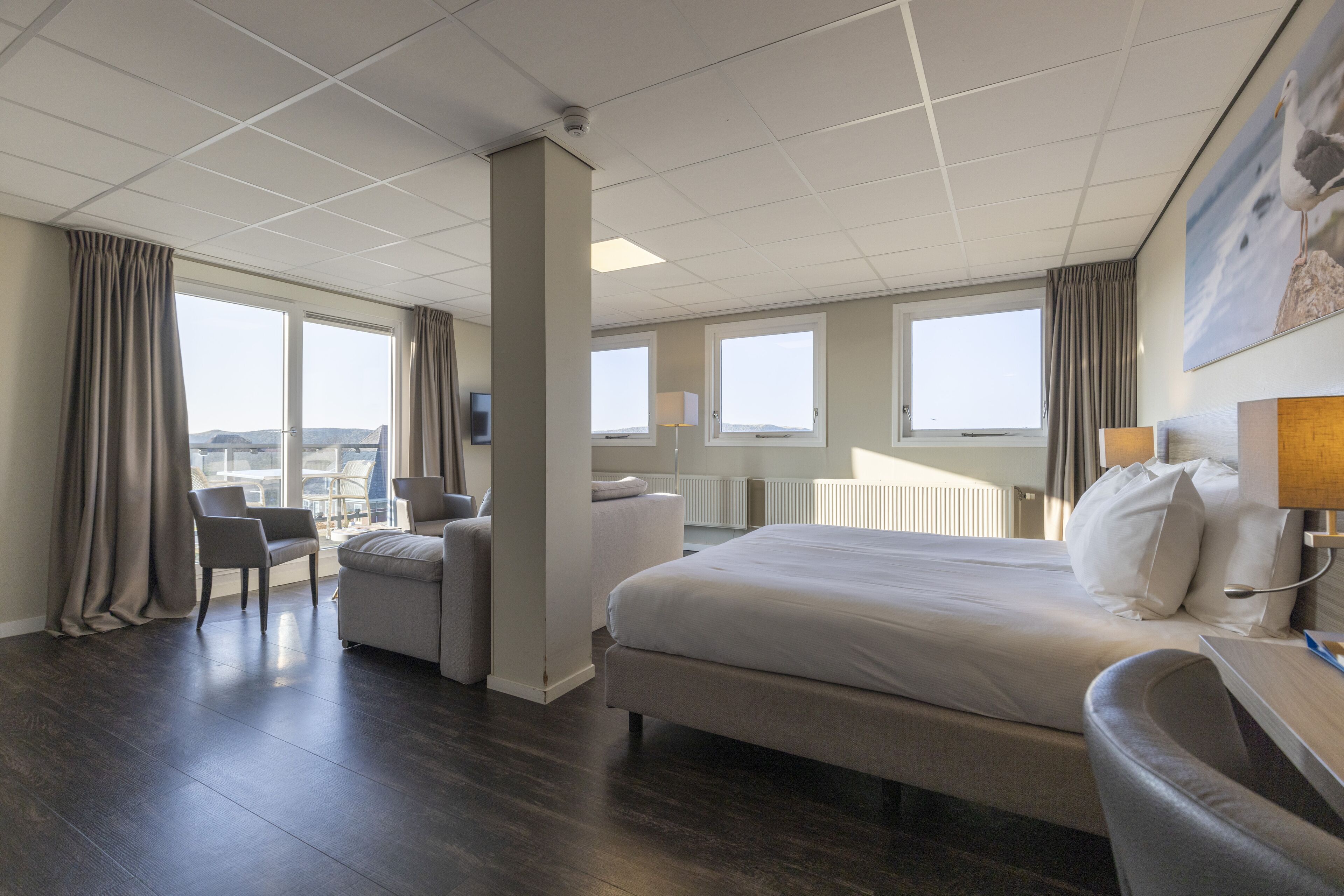 A hotel room with a large bed, a desk, a chair, and a view of the city through large windows.