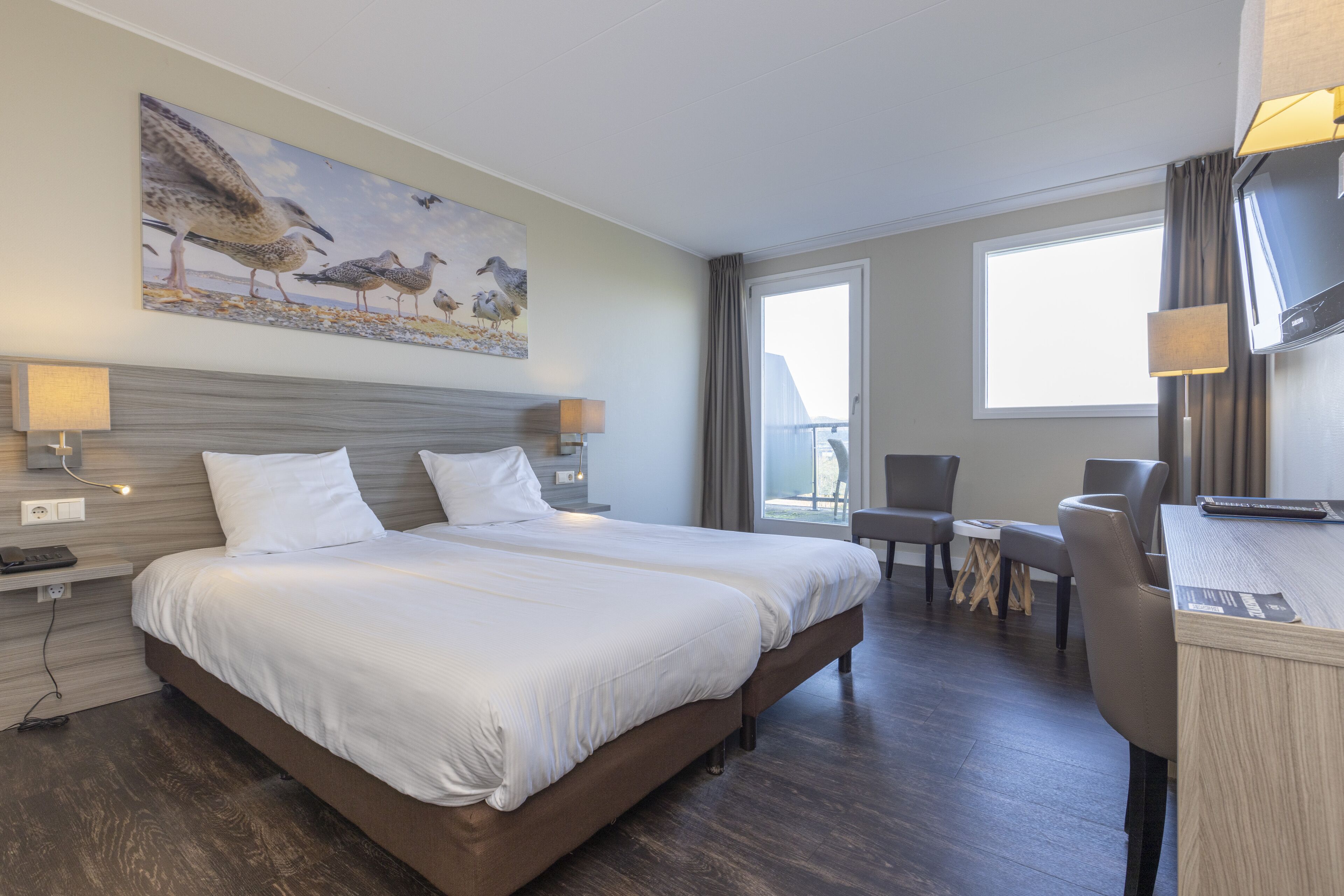 A hotel room with a large bed, two bedside tables, a desk with a chair, a painting of birds on the wall, and a window with curtains.