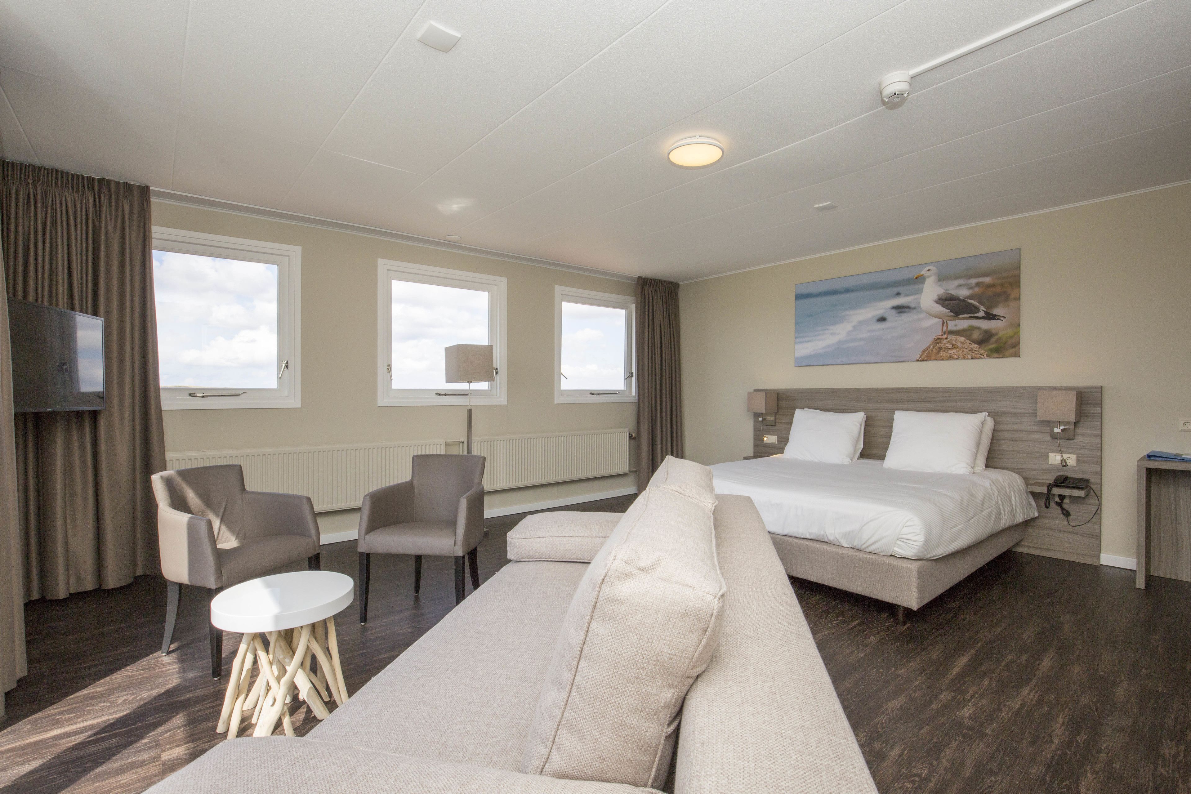 Una habitación de hotel moderna con una cama grande, dos sillones, un sofá, un televisor y un cuadro de una gaviota en la pared.