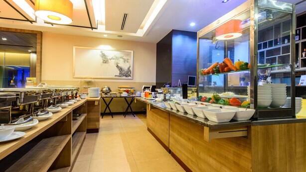 A modern buffet setup with multiple serving stations, a central island with a fruit display, and a large painting on the wall. - Mida Hotel Don Mueang Airport (Bangkok)