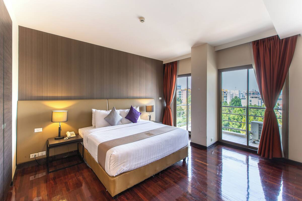 A hotel room with a large bed, a bedside table with a lamp, a telephone, and a view of the city through a large window.