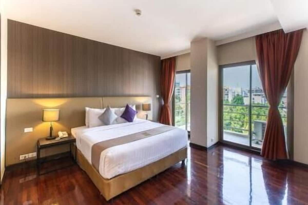 A hotel room with a large bed, a nightstand with a lamp, and a view of greenery through large windows.