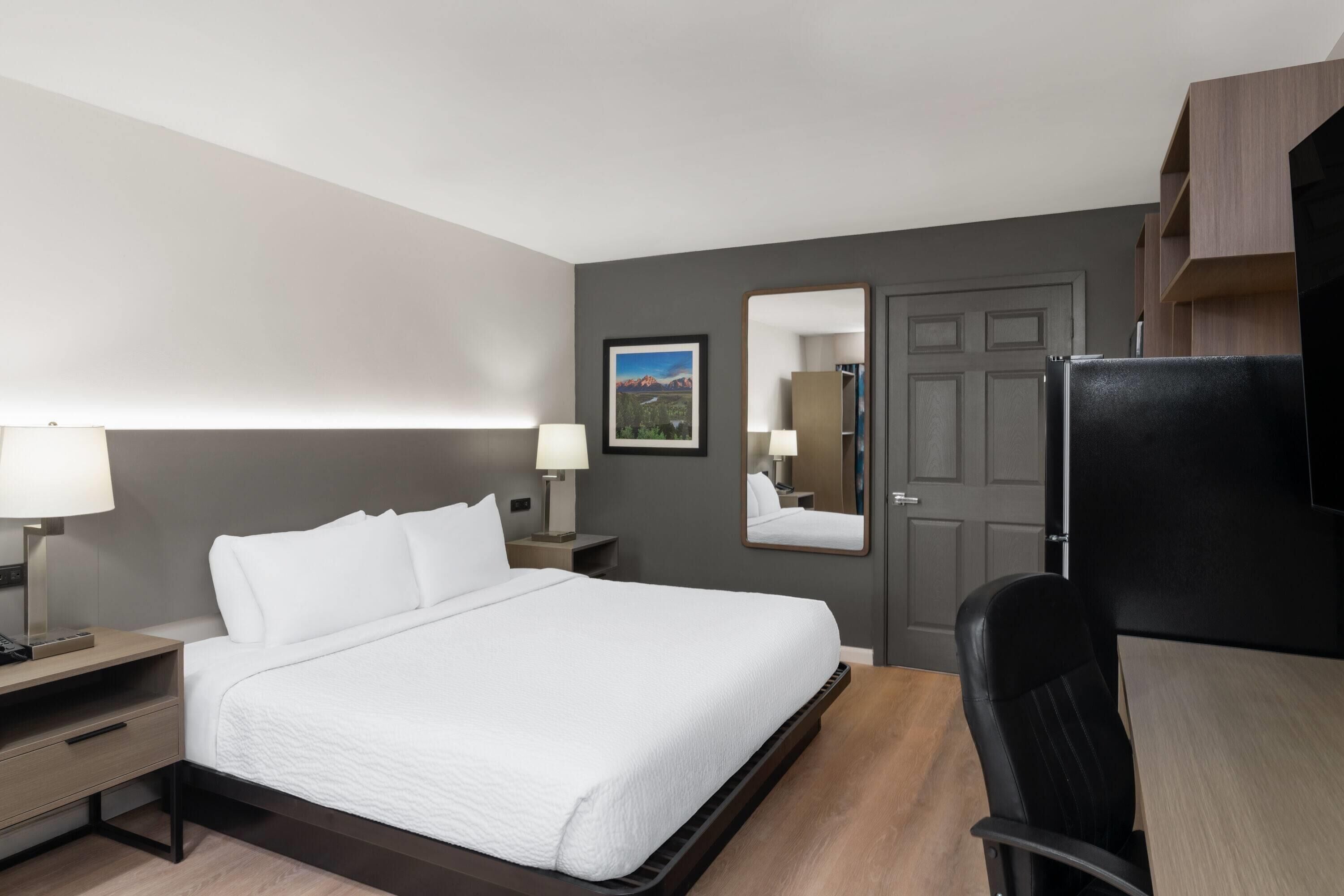 Una habitación de hotel con cama, mesitas de noche, escritorio, silla, refrigerador, espejo y puerta.