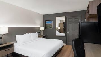 Una habitación de hotel con cama, mesitas de noche, escritorio, silla, refrigerador, espejo y puerta.