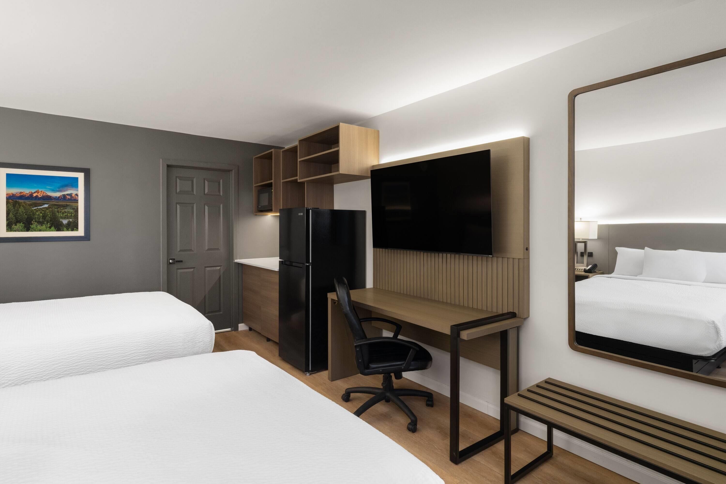 Une chambre d’hôtel avec deux lits, un bureau, une chaise, un réfrigérateur, une télévision et un grand miroir.